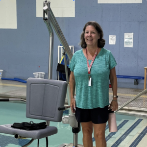 Photo of Kelly, Swimming Instructor at Worcester JCC. She is standing next to a gray motorized chair lift that helps people get in and out of the pool. She is wearing a turquoise short sleeved shirt and black shorts, as well as a red lanyard with a whistle.